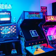 Neonowy napis SEGA nad starymi monitorami z grami. Obok automaty do gier z "Teenage Mutant Hero Turtles" i "Mortal Kombat", przypominające o czasach świetności emulatorów.